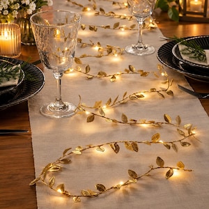 May include: A decorative string of lights featuring metallic gold-colored leaves intertwined with small, warm white LED bulbs. The flexible wire garland is arranged in a winding pattern across a light-colored fabric table runner between glassware.