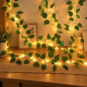 May include: A string of artificial green ivy leaves intertwined with warm white LED fairy lights. The vine features realistic heart-shaped foliage and thin flexible wire, creating a glowing decorative display that drapes across the surface.