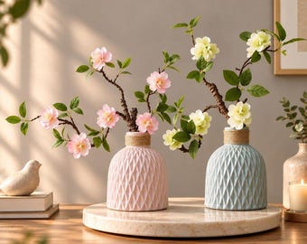 May include: Two ceramic vases with a geometric diamond pattern and a natural rope wrap around the neck. One vase is light pink and the other is light blue. Each holds branches with green leaves and delicate pink or pale yellow flowers.