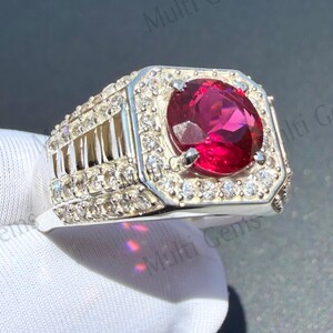 May include: A silver-toned ring featuring a large, round, deep red center stone held by four prongs. The rectangular head of the ring is surrounded by a border of small, clear, round-cut stones, with additional clear stones lining the band's sides.