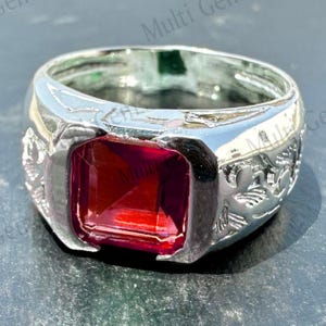 May include: A silver-toned ring featuring a large, square-cut, deep red gemstone set in the centre. The band of the ring displays an intricate, raised floral or vine-like pattern embossed along the sides, providing a textured metallic finish.