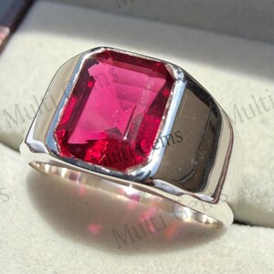 May include: A silver-toned ring featuring a large, rectangular, faceted deep red gemstone set in a smooth, polished metal band. The ring has a substantial, solid appearance with a clean, reflective surface surrounding the central stone.