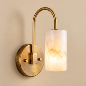 May include: A wall-mounted light fixture featuring a circular brass backplate and a curved arm that supports a cylindrical, translucent stone shade with a warm, marbled texture. The metal components have a brushed, aged brass finish.