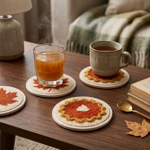 Op de afbeelding: Een set ronde, getextureerde punch needle onderzetters met herfstachtige ontwerpen, waaronder oranje esdoornbladeren en een pompoentaartmotief met crèmekleurige randen, geplaatst op een donkere houten tafel met twee gevulde drinkbekers.
