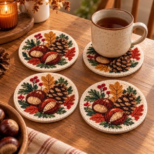 Op de afbeelding: Een set van vier ronde onderzetters met geborduurde herfstmotieven, waaronder bruine kastanjes, een donkerbruine dennenappel, groene dennennaalden, gele eikenbladeren en kleine rode bessen op een crèmekleurige stoffen ondergrond.