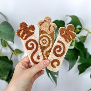 May include: Three rectangular punch needle bookmarks featuring textured yarn designs in shades of cream, tan, and brown. Each bookmark displays a unique swirl pattern and a scalloped top edge, crafted with a dense, looped fiber construction.