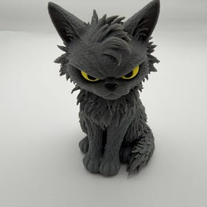 May include: A small, gray, sculpted figurine of a cat with a shaggy, textured coat and pointed ears. The cat has a stern facial expression with narrowed eyes and bright yellow irises, sitting in an upright, alert posture.