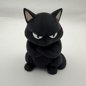 May include: A matte black cat figurine with a textured surface, pointed ears with light-colored inner details, and white eyes with black pupils. The cat is depicted in a seated position with its front paws crossed over its chest, displaying a stern facial expression with furrowed brows.