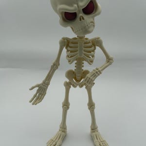 May include: A cream-colored articulated skeleton figurine with an oversized head and deep red eye sockets. The figure features a stylized, cartoonish skull with a prominent brow and a detailed rib cage, pelvis, and limb structure in a standing pose.
