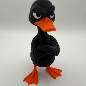 May include: A black rubber duck figurine with a textured surface, featuring a bright orange beak and webbed feet. The duck has a red crest on its head, white eyes with black pupils, and a stern facial expression with its wings crossed.