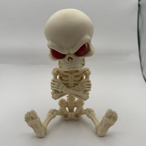 May include: A cream-colored, stylized skeleton figurine with a large, rounded cranium and deep-set, dark red eye sockets. The figure is depicted in a seated position with its arms crossed over its chest, featuring detailed rib cage and bone structures.