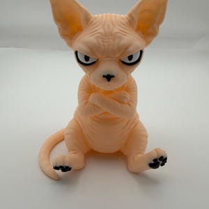 May include: A peach-colored Sphynx cat figurine with a wrinkled texture, large pointed ears, and a stern facial expression. The cat is depicted in a seated position with its arms crossed over its chest, featuring black eyes, a black nose, and black paw pads.