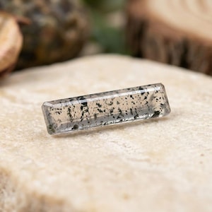 May include: A rectangular, clear glass or crystal bar featuring internal black speckled inclusions. The object has beveled edges and a smooth, polished surface, displaying a translucent appearance with scattered dark mineral-like flecks throughout.