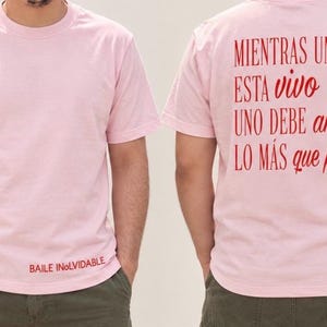May include: A light pink short-sleeved t-shirt featuring the red text "BAILE INOLVIDABLE" on the front bottom right and the red text "MIENTRAS UNO ESTA vivo UNO DEBE amar LO MÁS que pueda" printed on the back in a mix of serif and script fonts.