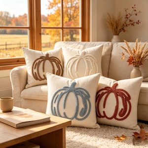 May include: Four square throw pillows with off-white fabric covers, each featuring a single raised, textured pumpkin design in a different color: brown, cream, dusty blue, and burnt orange, arranged on a light-colored sofa in a living space.