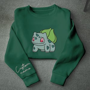 Op de afbeelding: Een donkergroene sweater met ronde hals en een centrale afbeelding van een lichtblauw wezen met rode ogen en een groene bol op de rug. De linkermouw bevat de witte cursieve tekst Custom name, geplaatst net boven de geribbelde boord.