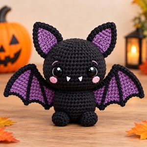 Peut inclure: Une peluche chauve-souris en crochet noir avec de grands yeux noirs, deux crocs blancs et des joues roses. La chauve-souris présente des intérieurs d'oreilles et des membranes d'ailes violets, un corps arrondi et de petits pieds.