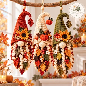 May include: Three hanging crochet gnome ornaments in autumn colors. The left gnome is rust-orange, the center is cream, and the right is olive green. Each features a sunflower, berries, leaves, wooden buttons, and a long, curly beard.