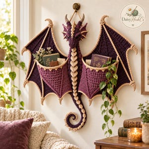 May include: A wall-mounted crochet dragon organizer in deep purple and cream yarn. The dragon features large wings with integrated pockets holding books and a trailing plant. A central spine and curled tail extend downward, with a logo in the corner.