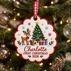 May include: A scalloped white fabric Christmas ornament featuring embroidered designs of a deer, a squirrel, a decorated Christmas tree, and gifts. The text reads Charlotte FIRST CHRISTMAS 2026. It hangs from a red and white gingham ribbon.