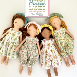 May include: Four handmade soft dolls with various skin tones, wearing floral and bunny-print dresses and jumpsuits. Each doll has a different hair color and a matching bow or headband. The dolls are arranged in front of a sign that reads "Prairie Lasses soft doll patterns".