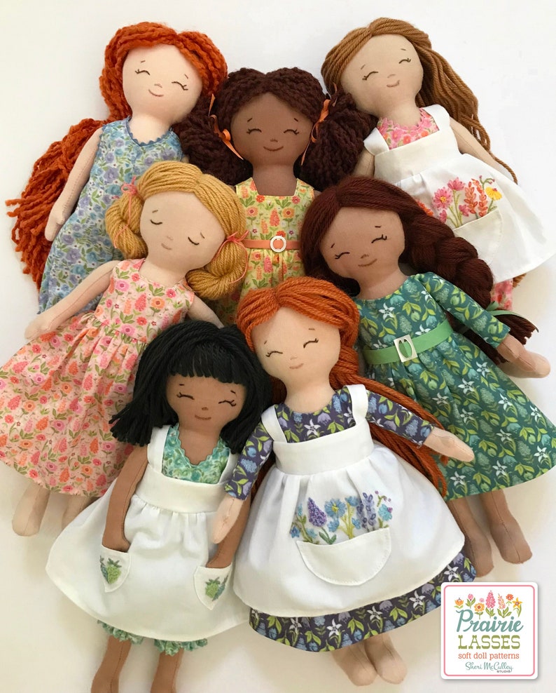 May include: Seven handmade fabric dolls with different hair colors and skin tones. Each doll is wearing a dress and an apron. The dolls are arranged in a circle, with their heads facing the center. The dolls are all smiling.