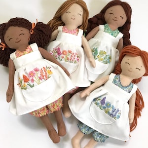 May include: Four handmade dolls wearing white pinafores with embroidered floral designs. Each doll has a different hair colour and skin tone. The pinafores have a large pocket on the front.