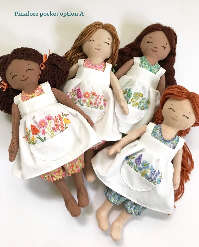 May include: Four handmade dolls wearing white pinafores with embroidered floral designs. Each doll has a different hair colour and skin tone. The pinafores have a large pocket on the front.