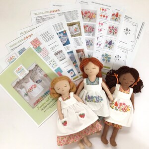 May include: Three handmade dolls with embroidered pinafores and bloomers. The dolls are dressed in white with floral embroidery. The dolls are lying on a white surface with printed instructions for making the dolls.