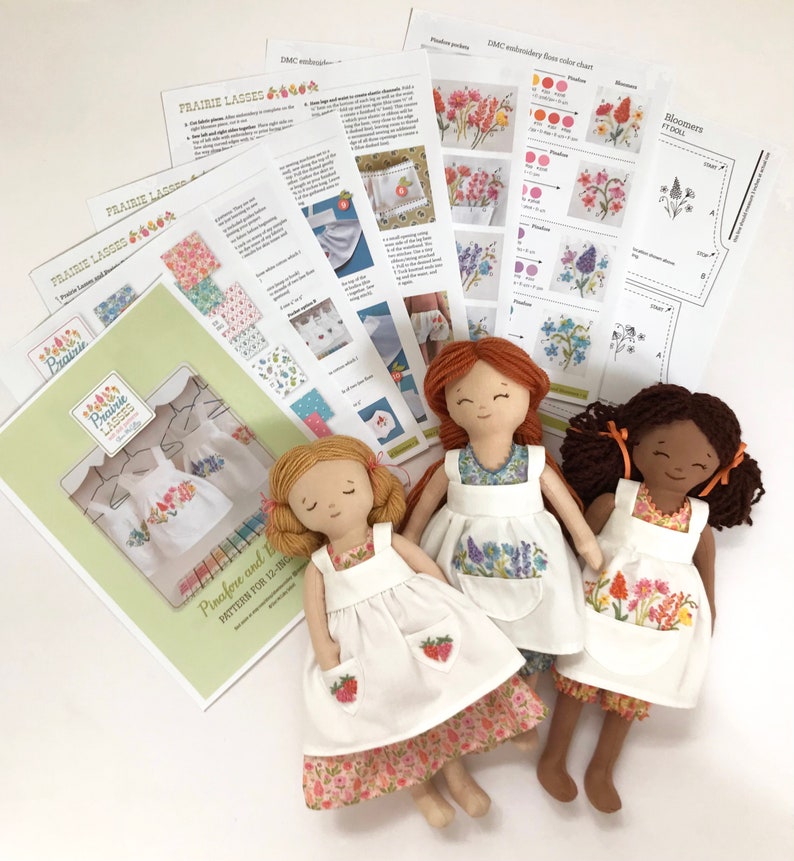 May include: Three handmade dolls with embroidered pinafores and bloomers. The dolls are dressed in white with floral embroidery. The dolls are lying on a white surface with printed instructions for making the dolls.