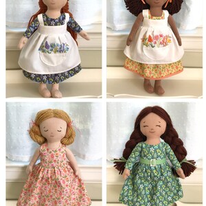 May include: Four handmade fabric dolls with different hair colors and clothing. Each doll is wearing a dress with a floral pattern. The dolls are standing on a white surface.