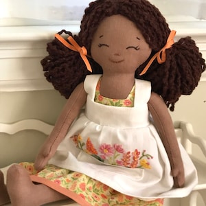 May include: A handmade doll with brown yarn hair tied with orange ribbons, wearing a white apron over a yellow floral dress.