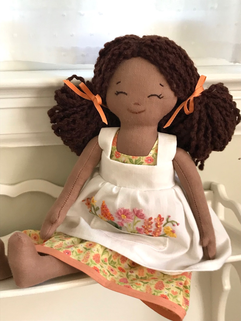 May include: A handmade doll with brown yarn hair tied with orange ribbons, wearing a white apron over a yellow floral dress.