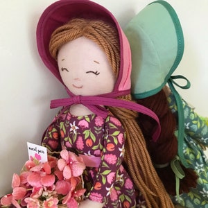 May include: Two handmade fabric dolls with long yarn hair. The doll on the left is wearing a pink and purple bonnet, a floral print dress, and holding a basket of pink flowers. The doll on the right is wearing a light green bonnet and a green and white floral print dress. The doll on the left has a tag that says "sweet pea".