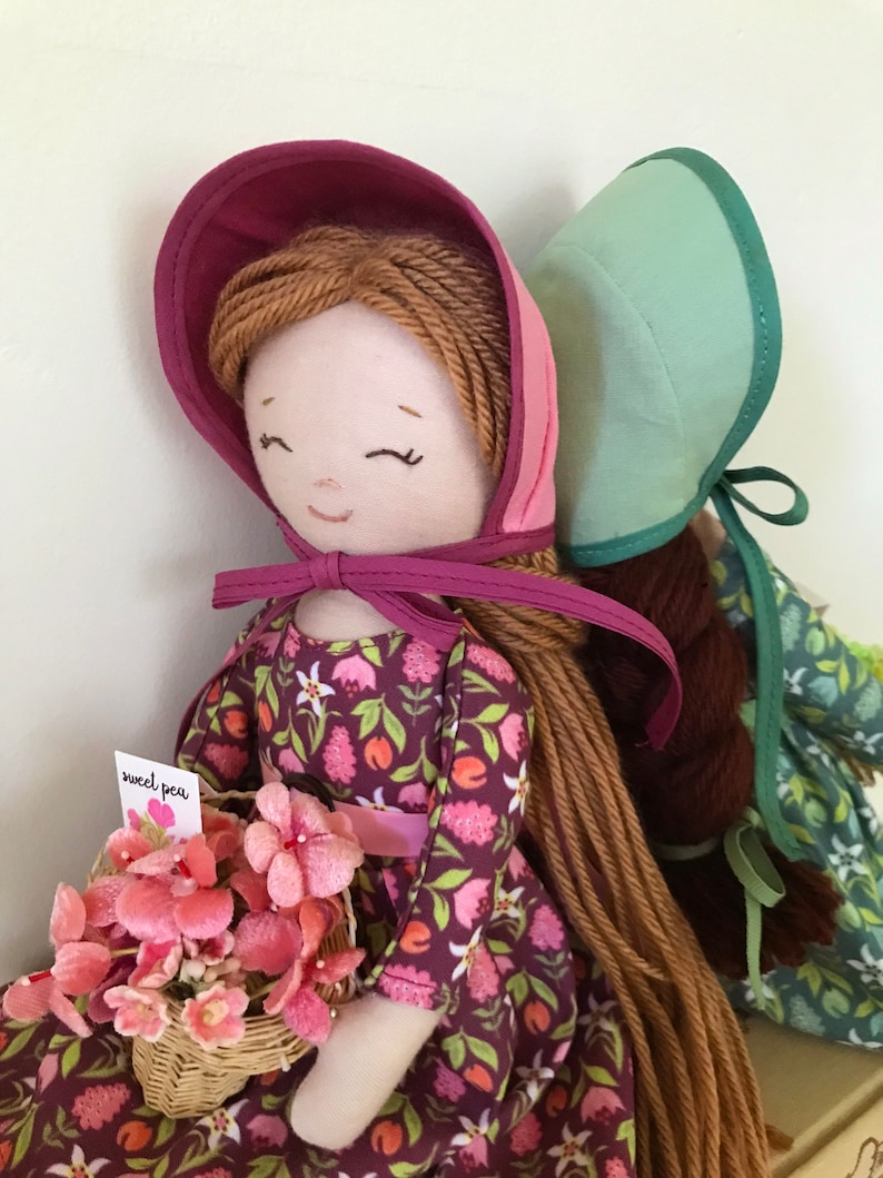 May include: Two handmade fabric dolls with long yarn hair. The doll on the left is wearing a pink and purple bonnet, a floral print dress, and holding a basket of pink flowers. The doll on the right is wearing a light green bonnet and a green and white floral print dress. The doll on the left has a tag that says "sweet pea".