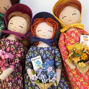 May include: Four handmade fabric dolls with embroidered faces and floral print dresses. Each doll holds a basket of flowers and a small card with the flower's name. The dolls are wearing bonnets in different colours: green, purple, blue, and yellow.