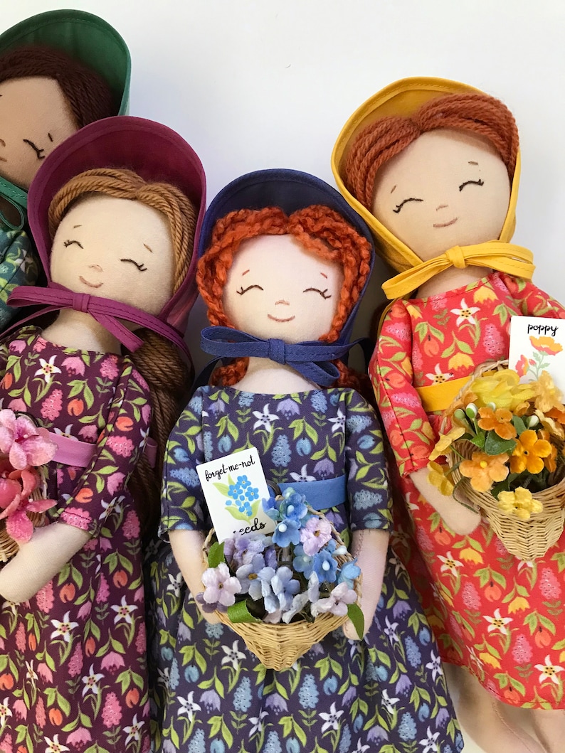 May include: Four handmade fabric dolls with embroidered faces and floral print dresses. Each doll holds a basket of flowers and a small card with the flower's name. The dolls are wearing bonnets in different colours: green, purple, blue, and yellow.