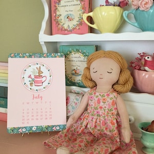 May include: A pink and green July calendar with a floral design and a rabbit illustration. The calendar is sitting on a table with a doll wearing a pink floral dress.