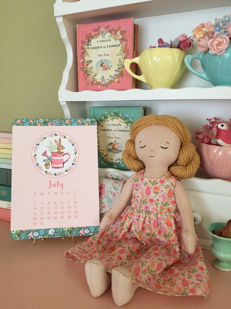 May include: A pink and green July calendar with a floral design and a rabbit illustration. The calendar is sitting on a table with a doll wearing a pink floral dress.