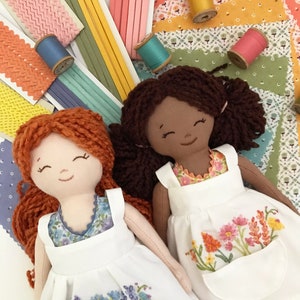 May include: Two handmade dolls with brown and red yarn hair wearing white dresses with embroidered floral designs. The dolls are sitting on a colorful fabric background with spools of thread.