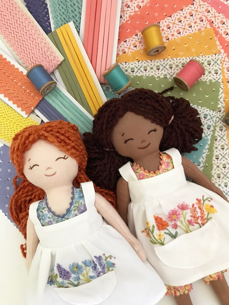 May include: Two handmade dolls with brown and red yarn hair wearing white dresses with embroidered floral designs. The dolls are sitting on a colorful fabric background with spools of thread.