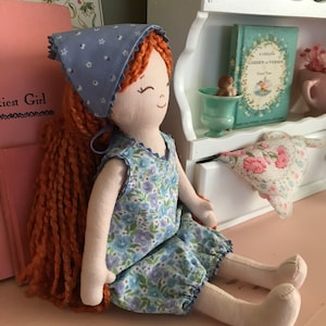 May include: A handmade rag doll with long, curly red hair wearing a blue and white floral dress and a blue bandana with white polka dots.