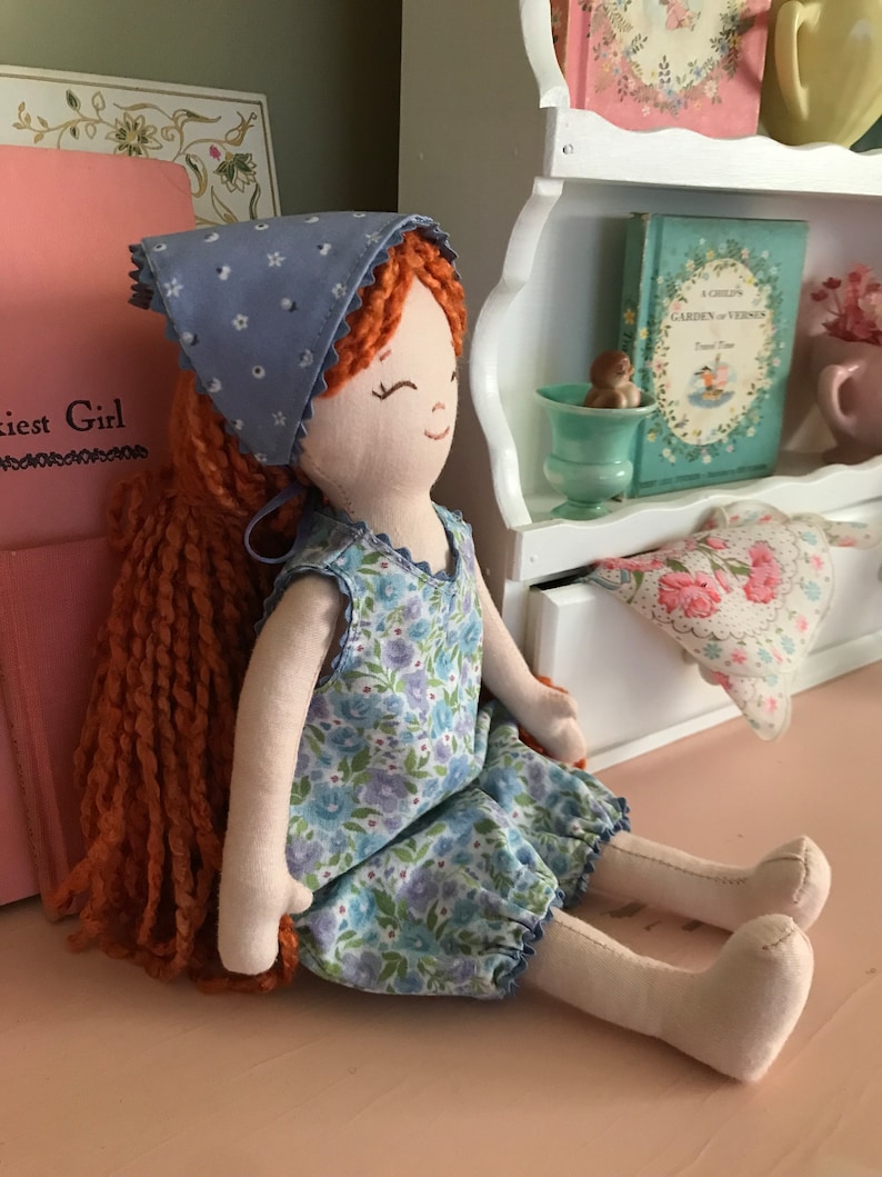 May include: A handmade rag doll with long, curly red hair wearing a blue and white floral dress and a blue bandana with white polka dots.