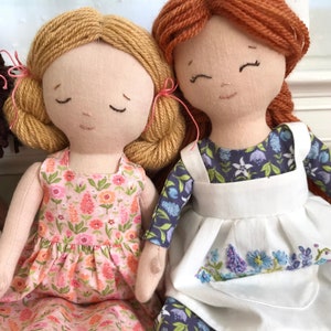 May include: Two handmade fabric dolls with yarn hair. The doll on the left has blonde yarn hair and is wearing a pink floral dress. The doll on the right has red yarn hair and is wearing a blue and white floral dress with a white apron.