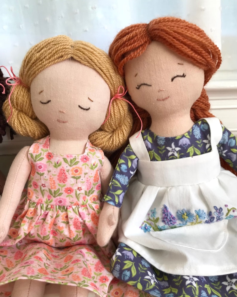 May include: Two handmade fabric dolls with yarn hair. The doll on the left has blonde yarn hair and is wearing a pink floral dress. The doll on the right has red yarn hair and is wearing a blue and white floral dress with a white apron.