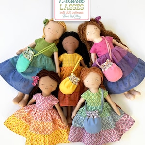 May include: Six soft fabric dolls with smiling faces and long hair, each wearing a colorful dress and carrying a small purse. The dresses are in shades of yellow, orange, pink, green, blue, and purple. The dolls are arranged around a sign that reads "Prairie Lasses."