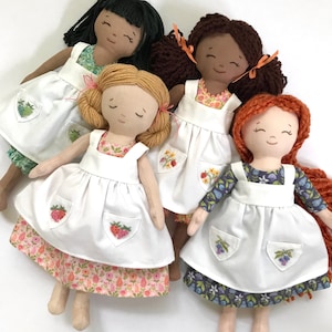 May include: Four handmade dolls wearing white pinafores with embroidered pockets. The dolls have different hair colours and skin tones. The pinafores have different floral patterns on the bottom.