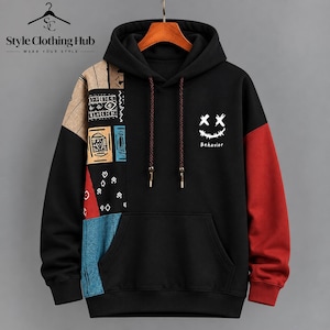 Puede incluir: Una sudadera con capucha negra con bolsillo tipo canguro y mangas largas. La manga derecha y el panel lateral muestran un diseño de retazos con patrones en beige, azul, rojo y negro. Un gráfico blanco muestra una cara sonriente cosida.