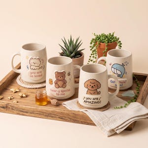 Può includere: Quattro tazze in ceramica maculata con illustrazioni a rilievo e testo. Una mostra un gatto con 'you are so loved', un orso con 'bear-y special to me', un cane con 'you are AMAZING!' e una balena con 'Get 'whale' soon!' stampati sopra.