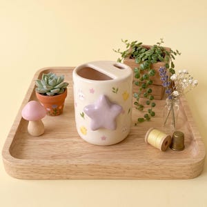 Puede incluir: Un soporte para cepillos de dientes de cerámica color crema con una estrella de color violeta claro en relieve en la parte frontal y pequeños motivos impresos de pollitos amarillos, conejos blancos, estrellas rosas y hojas verdes.